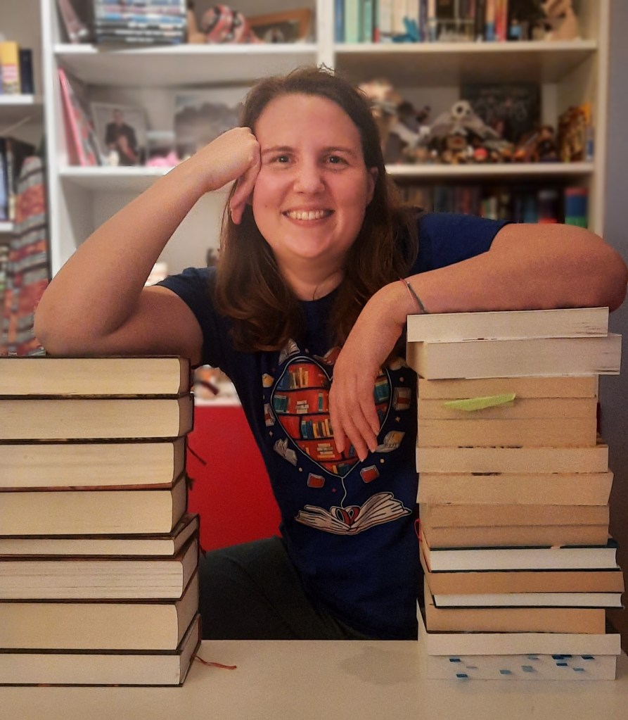 Eine Frau hockt vor einem Bücherregal, links und rechts von ihr ist je ein Bücherstapel, auf die sie sich mit ihren Ellbogen stützt. Sie lacht herzhaft.